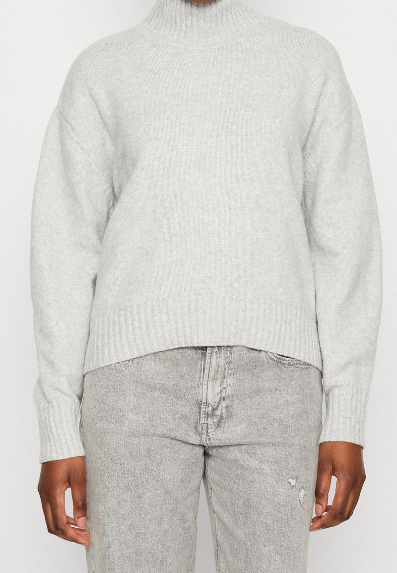 Pull gris clair en maille avec un col montant et des poignets côtelés. Porté avec un jean gris clair usé, présentant une coupe décontractée et une texture douce.