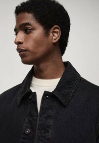 Dunkelblaue Jeansjacke mit Knopfverschluss und Kragen. Verfügt über goldene Nähte und schwarze Knöpfe. Trägt man über einem hellen Hemd.