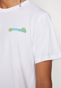 Camiseta de algodón blanca con un logotipo azul de "CONVERSE" y un gráfico de limón amarillo en el área del pecho izquierdo. Diseño de mangas cortas y cuello redondo.