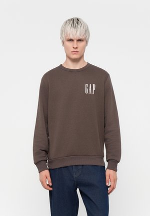 Felpa marrone con scollatura tonda, polsini e orlo a coste, caratterizzata da un logo bianco "GAP" sul lato sinistro del petto. Indossata con jeans blu.