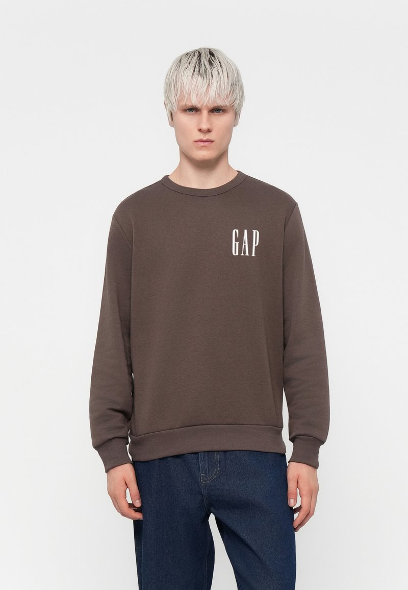 Brown sweatshirt with round neckline, ribbed cuffs, and hem, featuring a white "GAP" logo on the left chest. Worn with blue jeans.