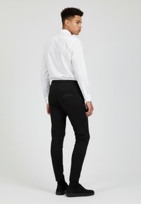 Schwarze, enganliegende Hosen mit glatter Textur, ausgestattet mit einer Tasche auf der Rückseite. Kombiniert mit einem weißen Langarmshirt. Minimales Design.