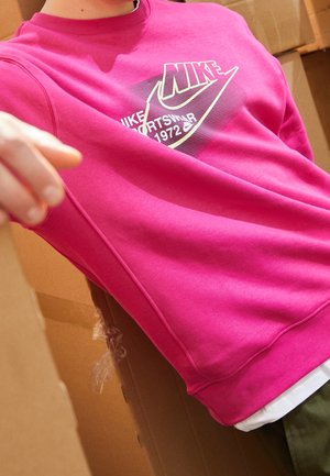 Person trägt ein knallpinkes Nike-Sweatshirt mit weißem und lila Logo-Schriftzug, umgeben von hohen braunen Kartons.