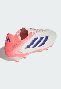 Botas de fútbol blancas con acentos en rosa y rayas azules, que presentan un material texturizado y un diseño curvado. Logo notable en el talón.