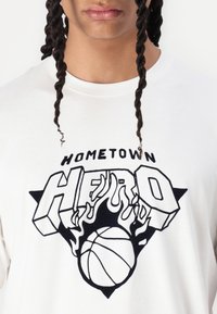 Hvid t-shirt med fed grafisk design i sort, der har teksten "HOMETOWN HERO", flammer og en basketball i midten.