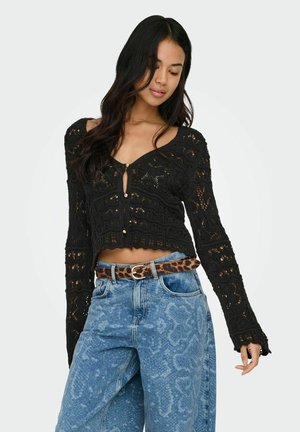 Femme portant un haut à manches longues en crochet noir et un jean taille haute bleu à motifs avec une ceinture imprimée léopard, debout devant un fond blanc.
