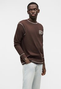 Καφέ μακρυμάνικο πουκάμισο με αντίθετες κρεμ ραφές και λογότυπο στο στήθος, συνδυασμένο με ανοιχτόχρωμο μπλε τζιν. Λείο βαμβακερό ύφασμα.