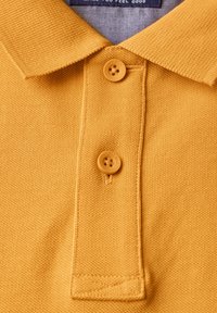 Żółta koszulka polo z teksturowanej tkaniny, z kołnierzykiem i dwoma guzikami w tym samym kolorze. Nić szycia jest widoczna w pobliżu guzików.