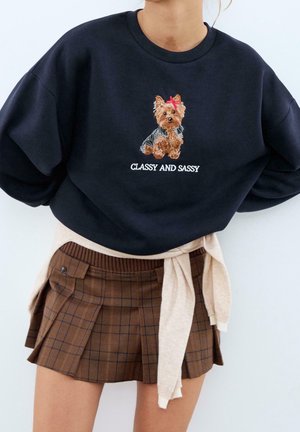 Frau trägt schwarzen Pullover mit gesticktem Hund und dem Text "Classy and Sassy", kombiniert mit braunem kariertem Faltenrock und beigefarbenem Pullover, der an der Taille gebunden ist.