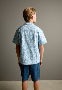 Kurzärmeliges blaues Hemd mit Smiley-Muster, kombiniert mit marineblauen Cargo-Shorts. Der Stoff wirkt leicht und der Schnitt lässig.