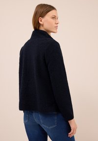 Ciemnoniebieska polarowa kurtka o teksturowanej powierzchni, z kołnierzem stojącym i długimi rękawami. Widok z tyłu ukazuje luźny krój i nieco skróconą długość.