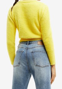 Suéter amarillo brillante y esponjoso con mangas largas y escote redondeado, combinado con jeans de mezclilla de tiro alto y lavado claro que cuentan con bolsillos traseros.