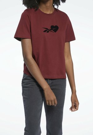 Person trägt ein dunkelrotes Kurzarm-T-Shirt mit einem schwarzen Herz und stilisiertem Design auf der Vorderseite, kombiniert mit dunkelgrauen Jeans, steht mit entspannten Armen.