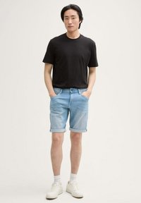 Schwarzes Baumwoll-T-Shirt, hellblaue Jeansshorts mit hochgerollten Säumen und weiße Sneakers, vor einem schlichten Hintergrund stehend.