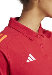Rotes Polohemd mit Kragen, mit orangenen Schulterstreifen und einem weißen Logo. Strukturierter Stoff mit zwei Knöpfen.