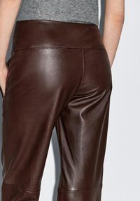 Bruine leren broek met een glanzende afwerking, voorzien van een brede tailleband en een gladde textuur. Het ontwerp is nauwsluitend met minimale naden.