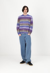 Multicolore gebreide trui met horizontale strepen in paars, blauw en bruin, ronde halslijn, gedragen over losse blauwe jeans en zwarte schoenen.