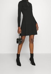 Robe noire à manches longues avec un col montant et un détail zippé, jupe évasée, en tissu texturé. Associée à des bottines noires et un mini sac à main.