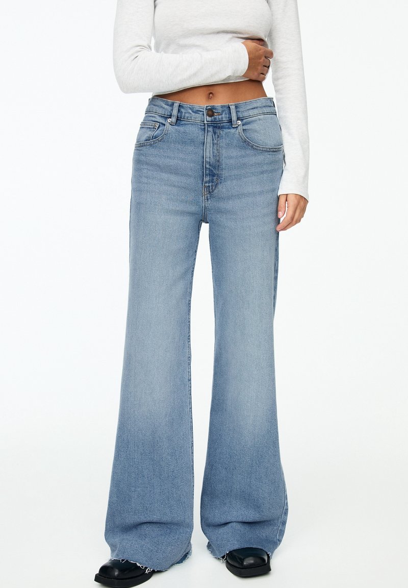 Femme portant un jean large bleu clair avec des ourlets effilochés, un crop top blanc à manches longues, et des chaussures noires à bouts carrés.