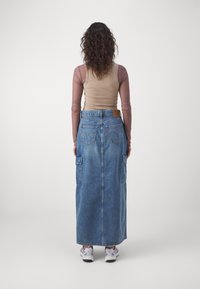 Denim maxi skirt with a straight cut, featuring two side pockets, five belt loops, and a leather tag at the back. Worn with a long-sleeve top.