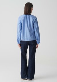 Camicia a maniche lunghe azzurro chiaro con yoke arricciato, abbinata a pantaloni in denim a gamba larga blu scuro, indossata con scarpe neutre.