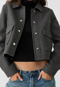 Gray wool cropped jacket with silver buttons, two front pockets, and a pointed collar, paired with a black ribbed top and high-waisted jeans.