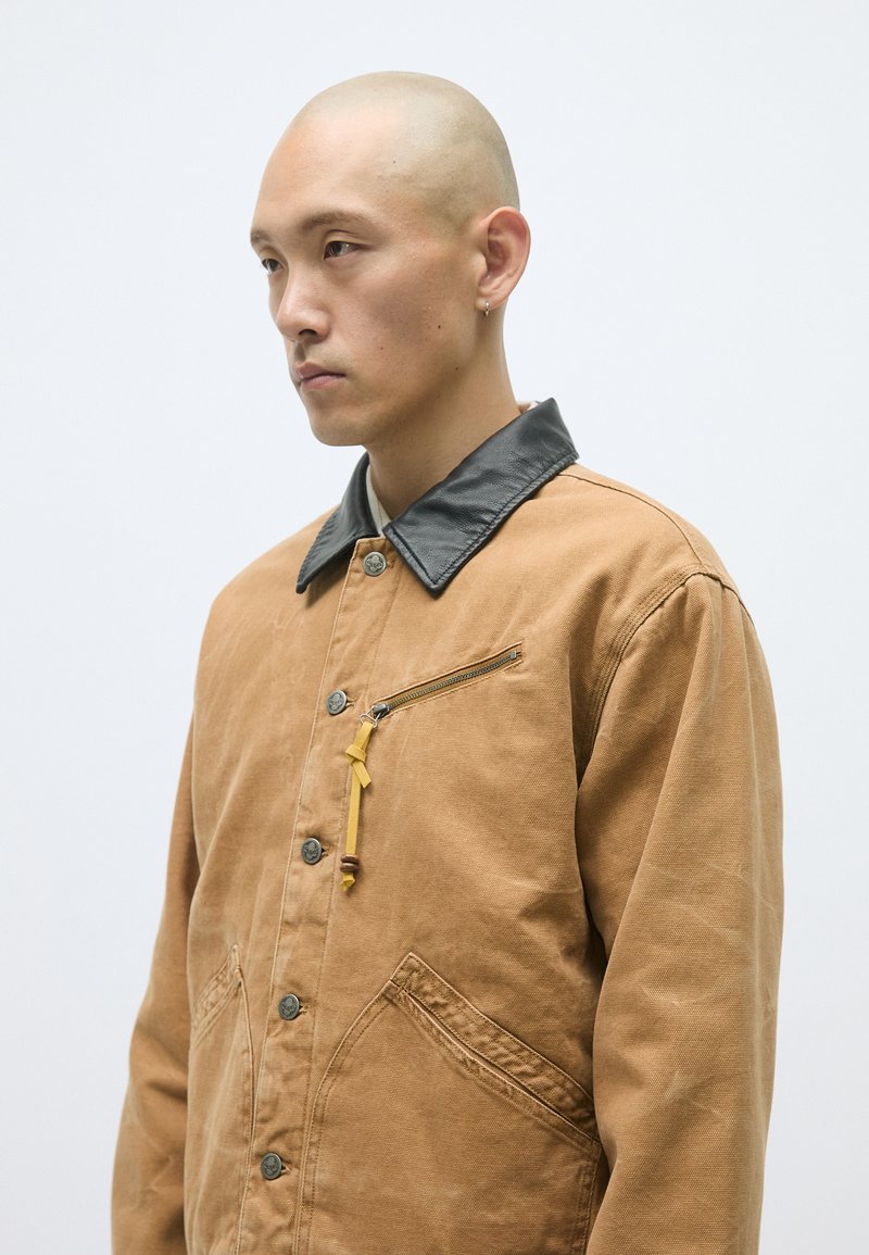 Braune Canvasjacke mit schwarzem Lederkragen, vorderer Reißverschlusstasche und gelben Akzenten. Mit Knopfverschluss und Seitentaschen.