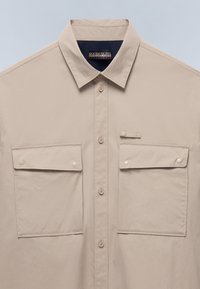 Camisa beige de botones con cuello clásico, dos bolsillos frontales y botones de presión blancos. Tejido suave y detalles de diseño sencillos.