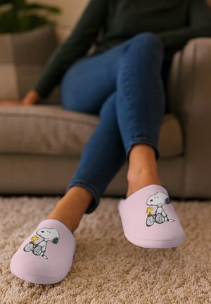 Persona seduta su un divano che indossa pantofole rosa con i personaggi Snoopy e Woodstock, gambe incrociate, su un pavimento beige carpet.
