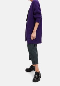 Lila stickad cardigan med ribbade ärmar, i kombination med mörkgröna avsmalnande byxor och kraftiga svarta skor. Har sidfickor.