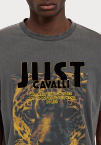 Pilkas medvilnės marškinėlis, apipavidalintas ryškiu juodu tekstu "JUST CAVALLI" ir spalvinga leopardų grafika. Suapvalintas iškirpimas ir laisvas siluetas su senoviniu look'u.