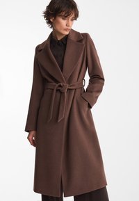 Brązowy płaszcz z paskiem, z klapami, długimi rękawami i prostym krojem. Wykonany z fakturowanej tkaniny, z przednim rozcięciem.