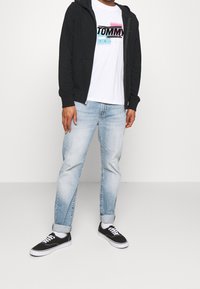 Sudadera negra con cremallera sobre una camiseta blanca con un logotipo gráfico de colores, jeans de mezclilla azul claro y zapatillas negras con suelas blancas.