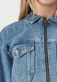 Light blue denim jacket with a zip front, pointed collar, and single chest pocket. Features visible stitching and textured fabric.