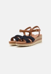 Navy suede sandals with brown leather straps, featuring a wedge base and a crisscross design. Light-colored sole with stitched detailing.