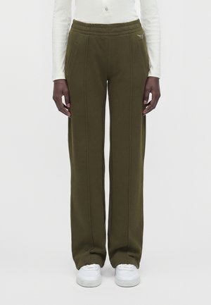 Personne portant un pantalon large vert olive avec coutures avant, un haut blanc à manches longues et des baskets blanches, debout devant un fond uni.