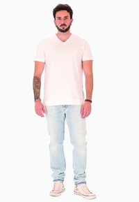 Uomo con capelli scuri e corti e barba, in piedi di fronte, indossa una t-shirt bianca a V, jeans azzurri chiari, scarpe da ginnastica bianche e un tatuaggio sul braccio sinistro.
