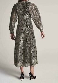 Robe longue en métal imprimé léopard noir et argent, avec des manches bouffantes et une silhouette fluide. Porte des talons noirs.