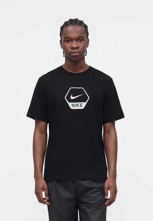 Νέος άνδρας με πλεξούδες, φορώντας μαύρο T-shirt Nike με εξαγωνικό λογότυπο και μαύρα σορτς, στέκεται μπροστά σε λευκό φόντο.