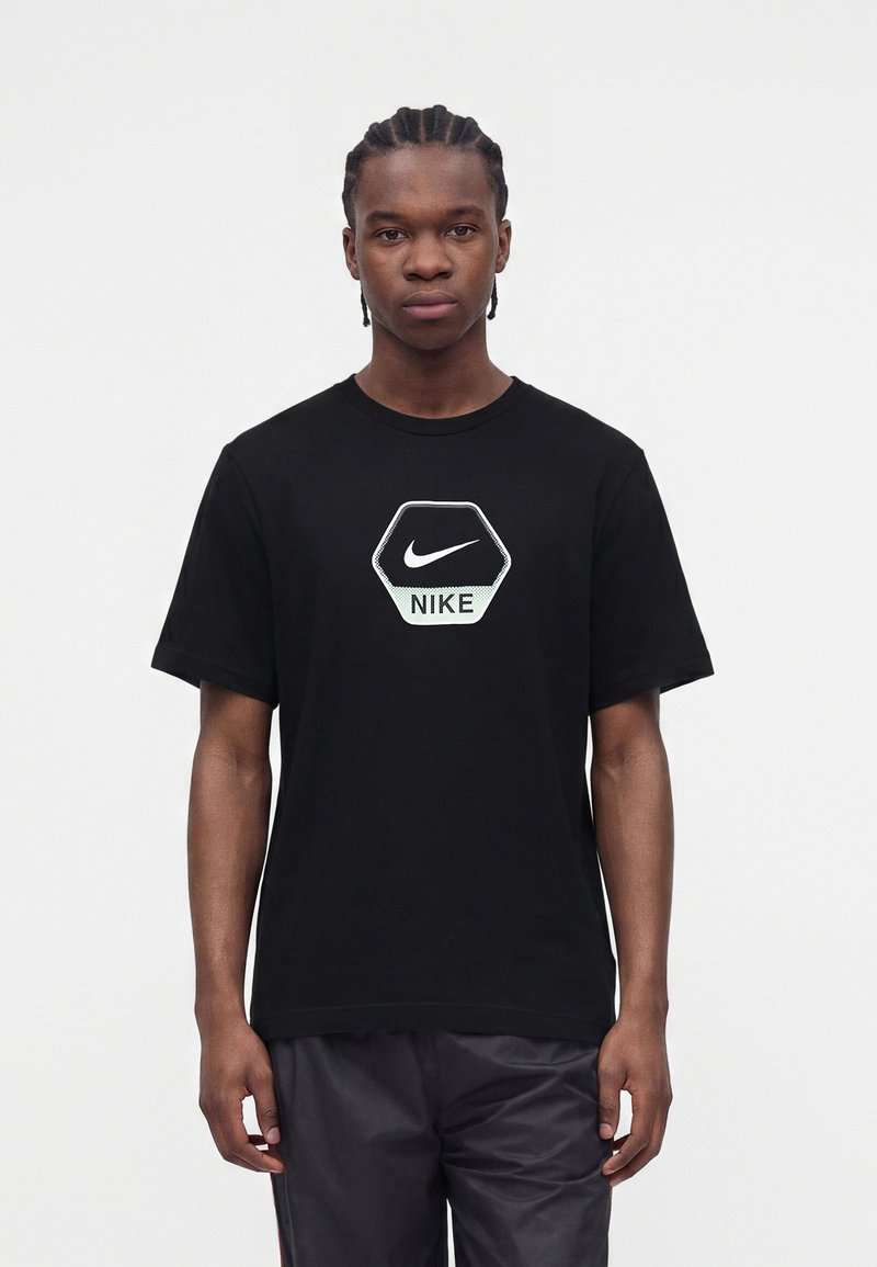 Νέος άνδρας με πλεξούδες, φορώντας μαύρο T-shirt Nike με εξαγωνικό λογότυπο και μαύρα σορτς, στέκεται μπροστά σε λευκό φόντο.