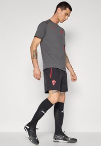 Sportstøj bestående af en grå trøje med røde detaljer, sorte shorts med sidelommer og logo, sorte knæhøje sokker og sorte sko.
