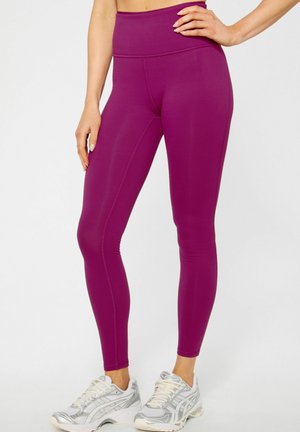 Woman wearing high-waist, plum-colored leggings and silver-white running shoes, standing with one hand on hip and the other relaxed.