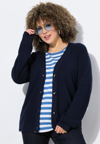 Cardigan en laine bleu marine avec un col en V, des boutons sur le devant et des poignets côtelés, porté par-dessus un haut rayé bleu et blanc. Des lunettes épaisses viennent rehausser le look.