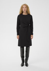 Czarna sukienka z długim rękawem i paskiem w talii, do długości połowy łydki, uszyta z gładkiego materiału. Połączona z czarnymi skórzanymi kozakami do kolan.