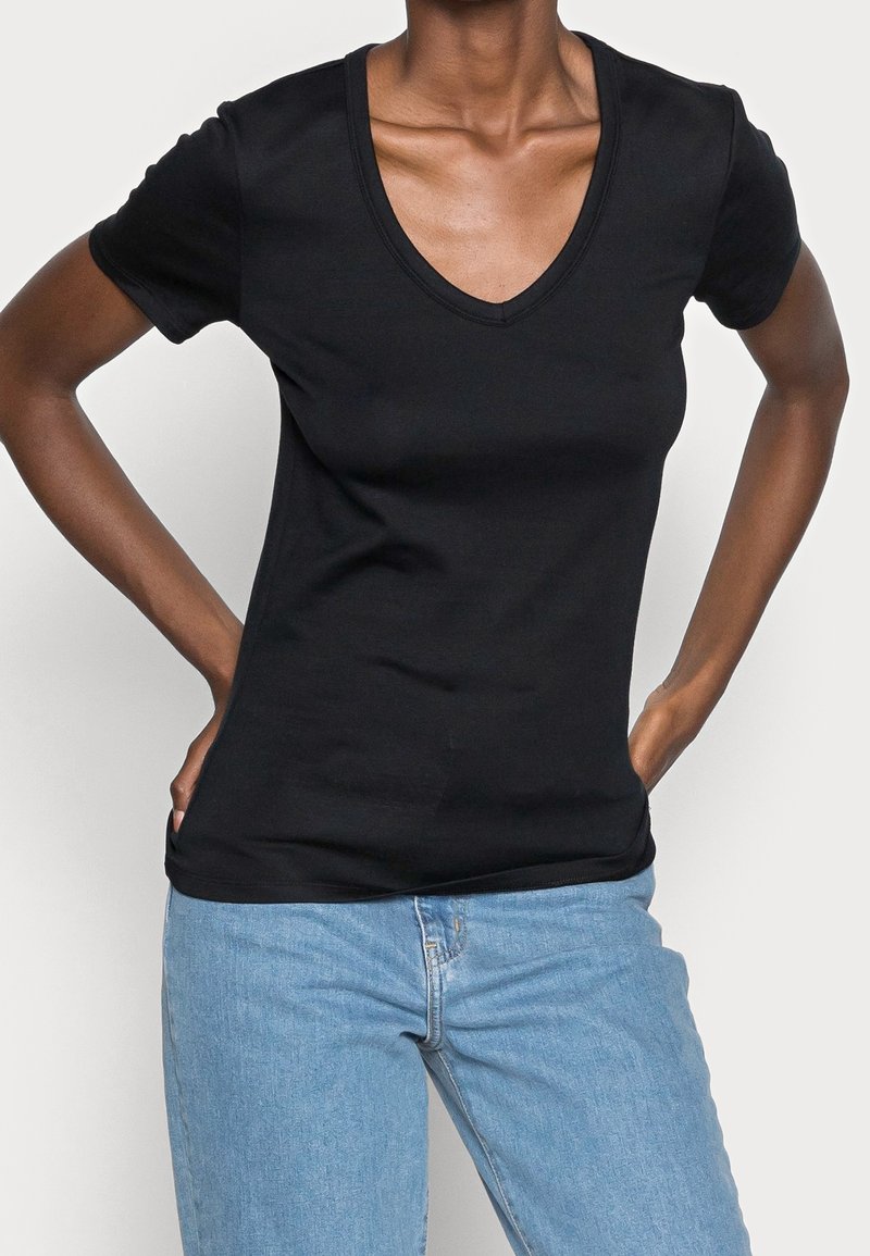 Personne portant un t-shirt noir col V à manches courtes et un jean bleu clair, mains posées sur les hanches devant un fond uni.