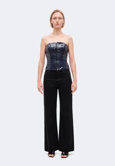 Un mannequin se tient devant un fond clair uni, vêtu d’un corset noir sans bretelles à la texture marquée, d’un pantalon à jambes larges et de sandales ouvertes à l’avant.