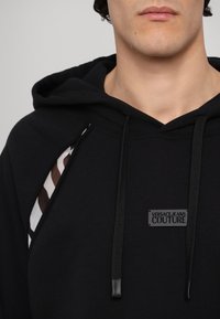 Černá mikina s kapucí s grafickým pruhem v hnědé a bílé barvě, s černou nášivkou, stahovací šňůrkou a strukturálním detailem látky.