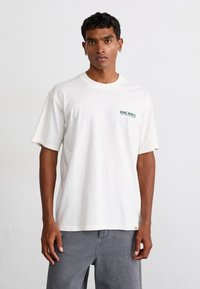 Tricou alb din bumbac, cu mâneci scurte, guler rotund și logo-ul verde "Dickies" pe piept. Modelul poartă pantaloni gri.