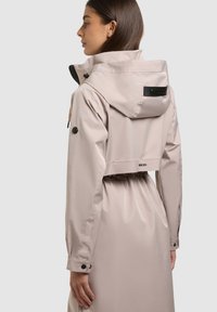 Ženska, ki nosi svetlo bež vodoodporno jakno z kapuco, nastavljivo pasom in zapognjenimi manšetami, obračena je vstran pred enobarvno ozadje.