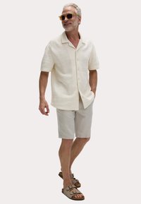 Chemise crème à manches courtes avec un motif texturé, associée à un short gris clair et des sandales marron. Comprend des lunettes de soleil en accessoire.